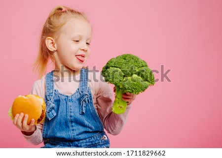 Hungry girl is holding a burger and broccoli in hands and show her tongue to broccoli on pink background. Unheathy vs healthy food. Concept of choice.