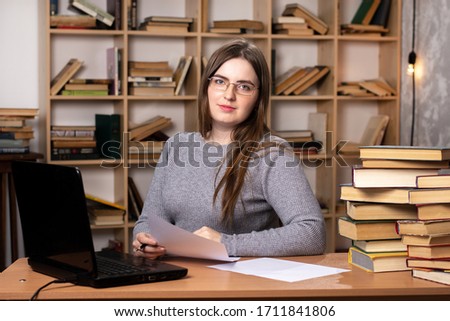 young girl in glasses sits at a desk with a laptop and books. large bookcase on the background. look at the camera with a smile
