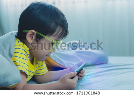 Adorable kid watching cartoon or playing game on smartphone. Young boy playing at bedroom. Lovely little boy wear eyeglasses when he was child because he always watching movie on mobile phone overtime