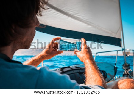 Adult male taking mobile phone pictures from the deck of a charter sail yacht with full sail on a sunny day with blue sky