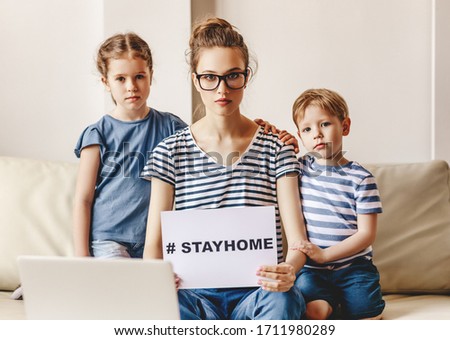 Serious young mother with little kids sitting in front of laptop on sofa and demonstrating paper with Stay Home hashtag for coronavirus prevention
