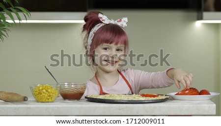 Cooking homemade pizza. Child girl adding sliced tomatoes to dough with sauce and cheese, smiles happy. Kid in kitchen dressed in apron and scarf like chef. Concept of nutrition, cooking, education