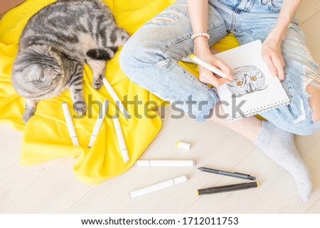 Drawing at home. A girl in blue jeans sketch a gray fold cat with yellow eyes. Nearby are markers, a yellow plaid. The cat is sitting near the girl. View from above, flat lay.