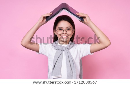 Funny Caucasian emotional brunette schoolgirl,student with glasses, wants to study by opening a book over her head in the form of a house, under protection, school, September 1, on a pink background.