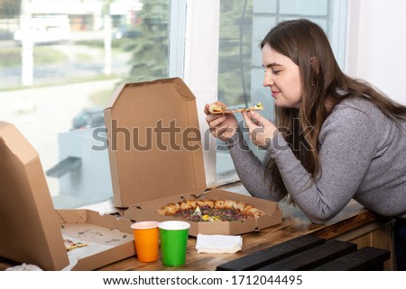 young girl holds a slice of pizza standing near the window. nearby are two paper boxes.