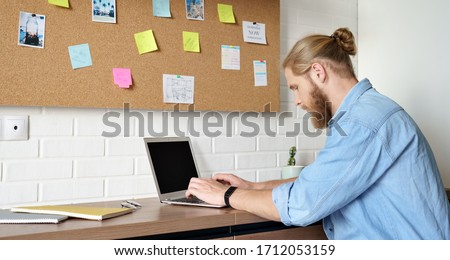 Serious young adult bearded casual man using laptop computer technology typing working or studying from home sitting at office desk. Millennial guy student elearning doing remote job in internet.