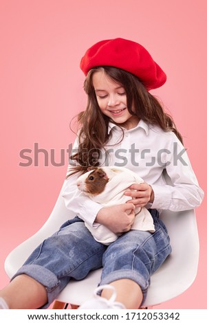 Cute happy little girl in stylish clothes holding guinea pig against pink background