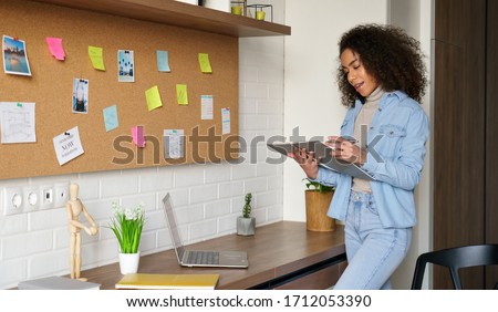 Smiling africanteen girl college student studying from home checking homework in notebook, elearning, stand at home office desk, making notes. Young black woman remote learning remote school lessons. Royalty-Free Stock Photo #1712053390