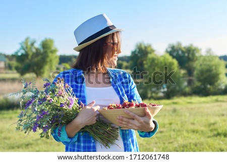 Female farmer with bowl of freshly picked strawberries and bouquet of wildflowers. Nature background, rural landscape, green meadow, country style