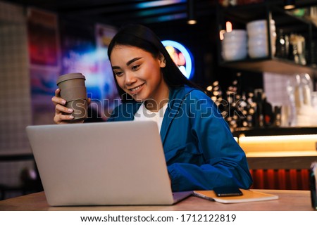 Photo of cheerful young asian woman working with laptop and drinking coffee while sitting in cafe