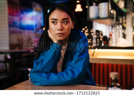 Photo of beautiful young asian woman posing and looking aside while sitting in cafe