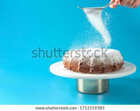 Woman's hand sprinkling icing sugar over fresh home made bundt cake. Powder sugar falls on fresh perfect bunt cake over blue background. Copy space for text. Ideas and recipes for breakfast or dessert Royalty-Free Stock Photo #1712159383