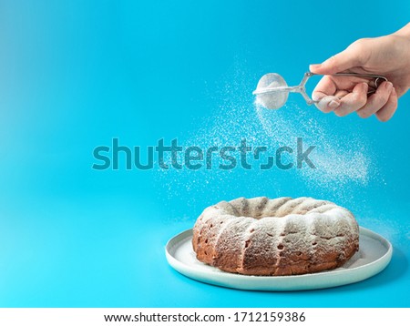 Woman's hand sprinkling icing sugar over fresh home made bundt cake. Powder sugar falls on fresh perfect bunt cake over blue background. Copy space for text. Ideas and recipes for breakfast or dessert Royalty-Free Stock Photo #1712159386