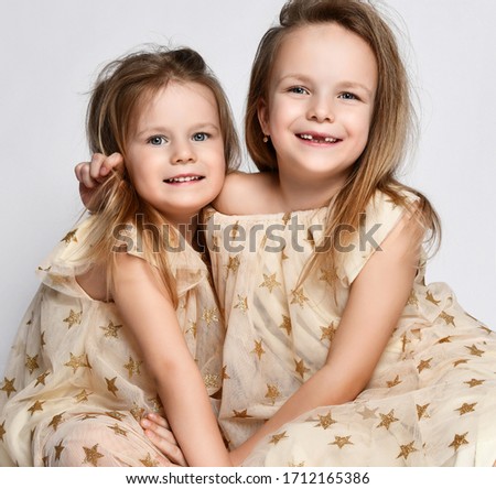 Portrait of two small beautiful smiling girls sisters in same dresses with stars sitting on floor and cuddling over grey background. Happy childhood, stylish home children clothes concept