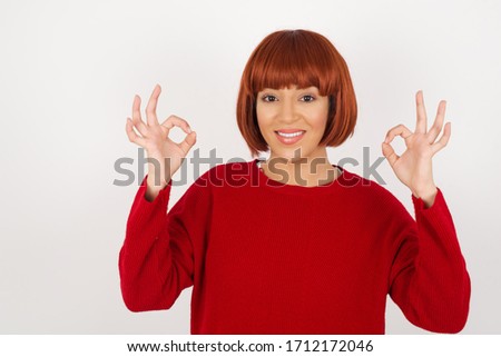 Glad attractive woman shows ok sign with both hands as expresses approval, has cheerful expression. Photo of beautiful female has appealing appearance, being optimistic. Standing against gray wall.