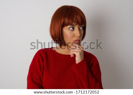 Shot of contemplative thoughtful Young woman keeps hand under chin, looks thoughtfully upwards, dressed in casual clothes, poses over white background with free space for your text