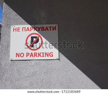  Car parking sign. The red  inscription in ukrainian and in english  "No parking." White rectangle on a gray rough wall. A shadow divides the frame obliquely.