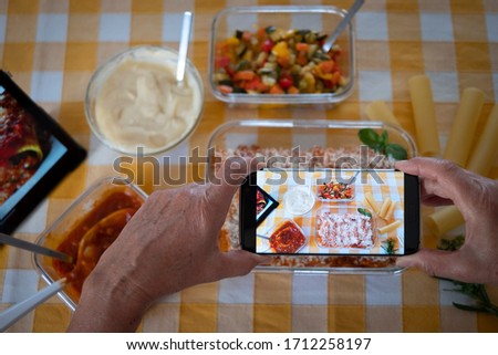 The hands of an elderly woman take a picture of the pan full of cannelloni stuffed with vegetables and ragù. Handmade. Following the Italian recipe on the tablet