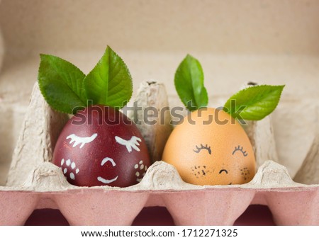 two chicken colored eggs with a cute painted face on them, lying in an egg box on a pink background, a festive Easter concept