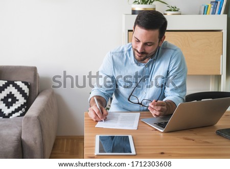 Young businessman sitting at a table working at home on a laptop and writing on paper stock photo