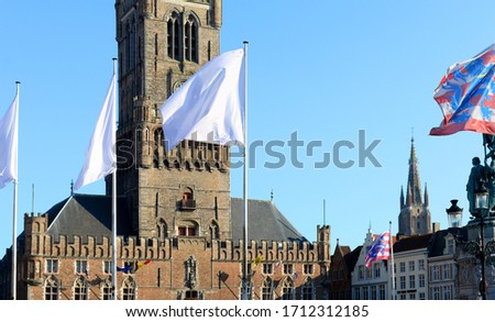 White flags, symbol of solidarity, and flags of Bruges in front of belfry with church of Our Lady in the background, against blue sky