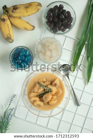 top view.  Banana compote in a large bowl and three colors of palm sugar fruits in three small bowls.  three bananas and two pandan leaves on a white table.  selective focus.