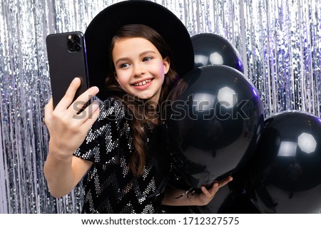 Portrait of a narcissistic teenager on a birthday taking selfie on a smartphone among black helium balloons on a shiny background
