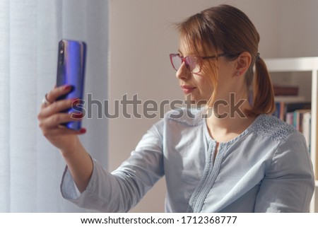 Young adult caucasian woman female girl sitting in the chair at home or work in front of the bookshelf holding smartphone mobile phone taking selfie photos or making a video call in day side view