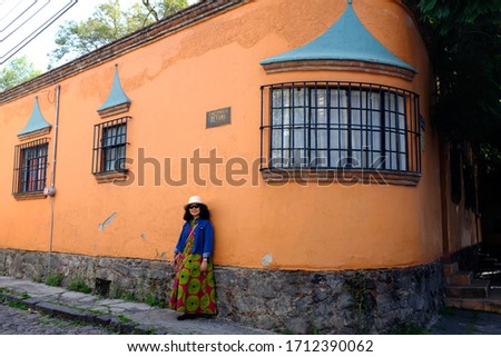 An Asian female tourist posing for a picture against an old bright orange building in Coyoacán neighbourhood, Mexico City, Mexico. The sign says “Rinconada Retama” which is a street name.