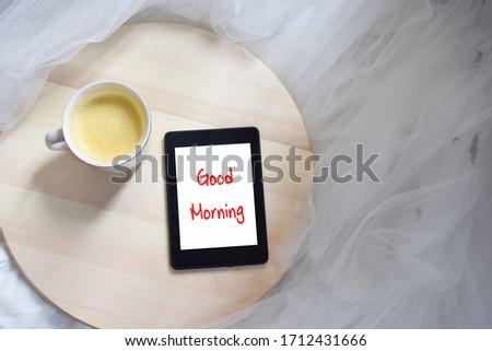 Flat lay of a tablet and a cup of coffee on a circular wooden table laid on a white tulle fabric. Words good morning printed on a tablet.