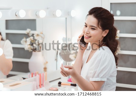 Morning of beautiful young woman applying makeup and talking by phone at home