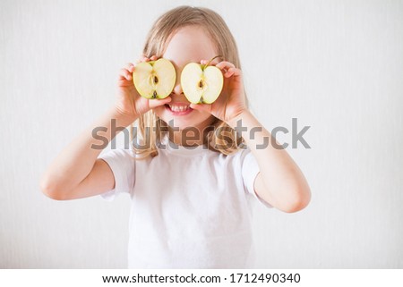 A cheerful little blonde girl smiling, having fun and looking through two halves of an apple in front of her eyes. Fruit. Symbol of the Jewish New Year. Rosh ha Shana.