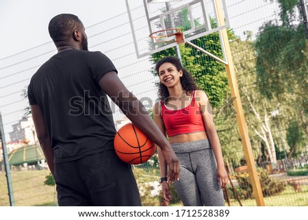 Young african descent couple standing on basketball court outdoors man back view holding ball looking at woman walking towards laughing playful