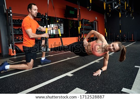 Coach motivating and training young woman smiling motivated doing side-plank tap TRX exercise at gym using special equipment haging sporty lifestyle