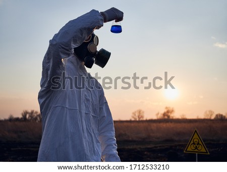 Scientist in white coverall, gas mask and gloves holding test tube with blue liquid while studying soil samples on scorched territory with biohazard sign, doing research work in field with burnt grass