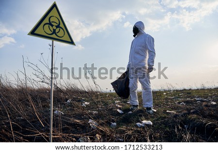 Environmentalist in protective suit and gas mask holding garbage bag, standing near biohazard sign on abandoned territory with trash, looking away. Concept of ecology and biological hazard. Royalty-Free Stock Photo #1712533213