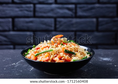Black bowl with asian fried jasmine rice with shrimps scrambled eggs and spring onions with thai sauce on a dark concrete background in front of black brick wall, close-up, copy space Royalty-Free Stock Photo #1712560408