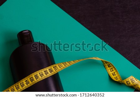 Black plastic bottle and tailor meter on a turquoise background. Black bottle and yellow ribbon of a tailor's ruler. Side view. Close-up. Selective focus. Free space for designer text.