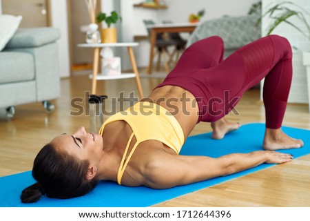 Young athletic woman lifting her hips while doing glute bridge exercise on the floor at home.  Royalty-Free Stock Photo #1712644396