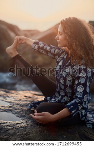 Beautiful curly woman by the ocean. Woman yoga portrait. Breathtaking sunset. Ocean splashes. Amazing graceful woman doing yoga by the ocean sunset. Yoga poses.
