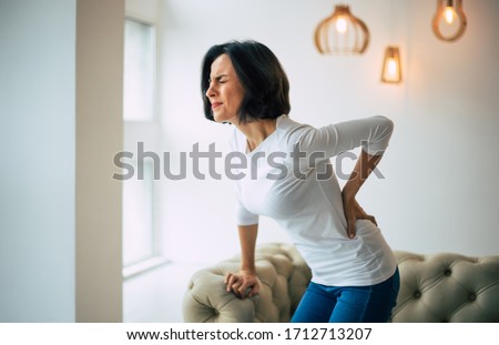 Chronic back pain. Adult woman is holding her lower back, while standing and suffering from unbearable pain. Royalty-Free Stock Photo #1712713207