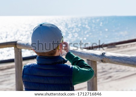 Side view of Young boy in casual clothes wearing face mask taking photos with his smartphone on beach. Mobile photographer taking photos on the shore.