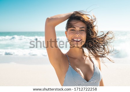 Young smiling woman in bikini at tropical beach looking at camera. Portrait of beautiful latin girl in swimwear with ocean in background. Happy tanned hispanic woman relaxing at sea and copy space.
