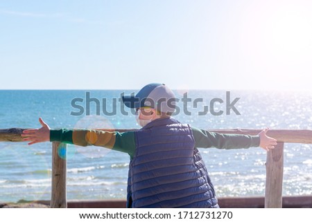  caucasian boy in casual clothes at the seaside pebble beach on overview ramp outstretched hands looking to the side