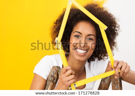 Excited African American woman in her new home holding up a wooden frame in the shape of a house to celebrate her achievement as she stands in front of a half painted wall