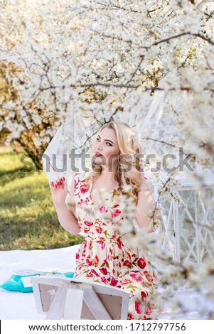 Positive,pretty,blond woman artist painting a picture under full white blooming trees.Nature inspiration for painting,drawing with easel, brush.Spring photoshoot in the apple garden in sunset,outdoor.