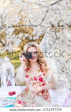 Positive,pretty,blond woman with red lips take photos by old retro camera under full white blooming trees.Spring photoshoot in the apple garden,outdoor.Portrait of beautiful smiling photographer.
