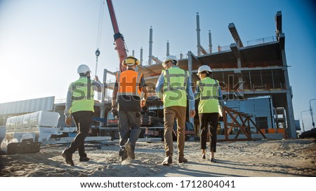 Diverse Team of Specialists Inspect Commercial, Industrial Building Construction Site. Real Estate Project with Civil Engineer, Investor and Worker. In the Background Crane, Skyscraper Formwork Frames Royalty-Free Stock Photo #1712804041