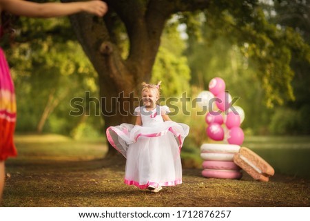 Little blond pretty girl with tails in white long party dress holding birthday cake with candles and dancing on the grass summer glade near balloons and macaroons