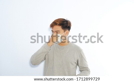 Image of pretty asian man standing against white wall with mask on his mouth dressed in long grey shirt. He shut his mouth up to prevent corona virus COVID-19. Sickness, illness, healthcare